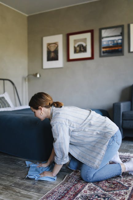 A woman with curly hair, wearing a beige jacket, dark jeans, and white shoes, is engaged in surface cleaning inside a living room. She is using a yellow vacuum cleaner with a long black hose and a wid
