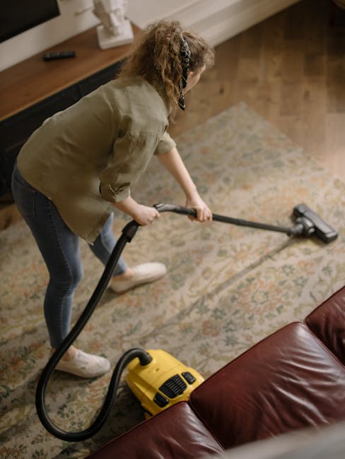 A woman with auburn hair tied back, wearing a light blue and white striped shirt and blue jeans, is kneeling on an ornate, multicolored rug in a living room. She is using a blue cloth to wipe and dust
